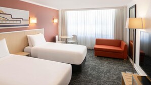 Ein Hotelzimmer mit zwei Betten, einem kleinen Tisch, einer Couch, einem Fernseher und einem Fenster mit Vorhängen.