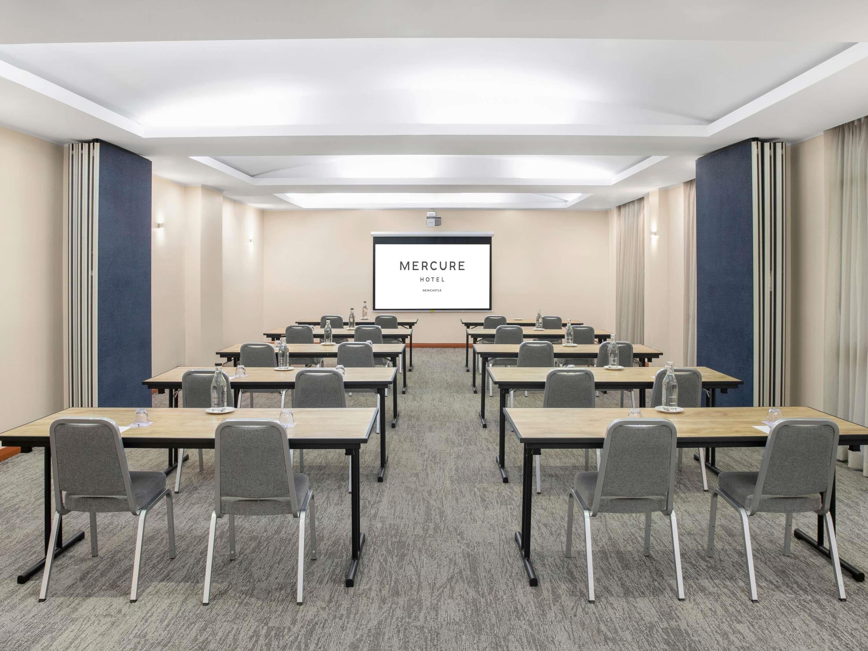 Un salón de conferencias con una pantalla grande que muestra 'Mercure Hotel Newcastle', filas de mesas con sillas y suelo alfombrado.