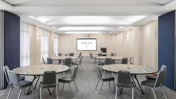 A conference room with multiple tables and chairs, a projector screen displaying 'Mercure Hotel Newcastle', and a carpeted floor.