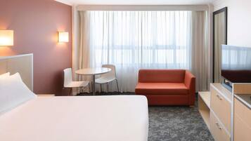 Habitación de hotel con cama, sofá, televisión, escritorio con sillas y ventana con cortinas.