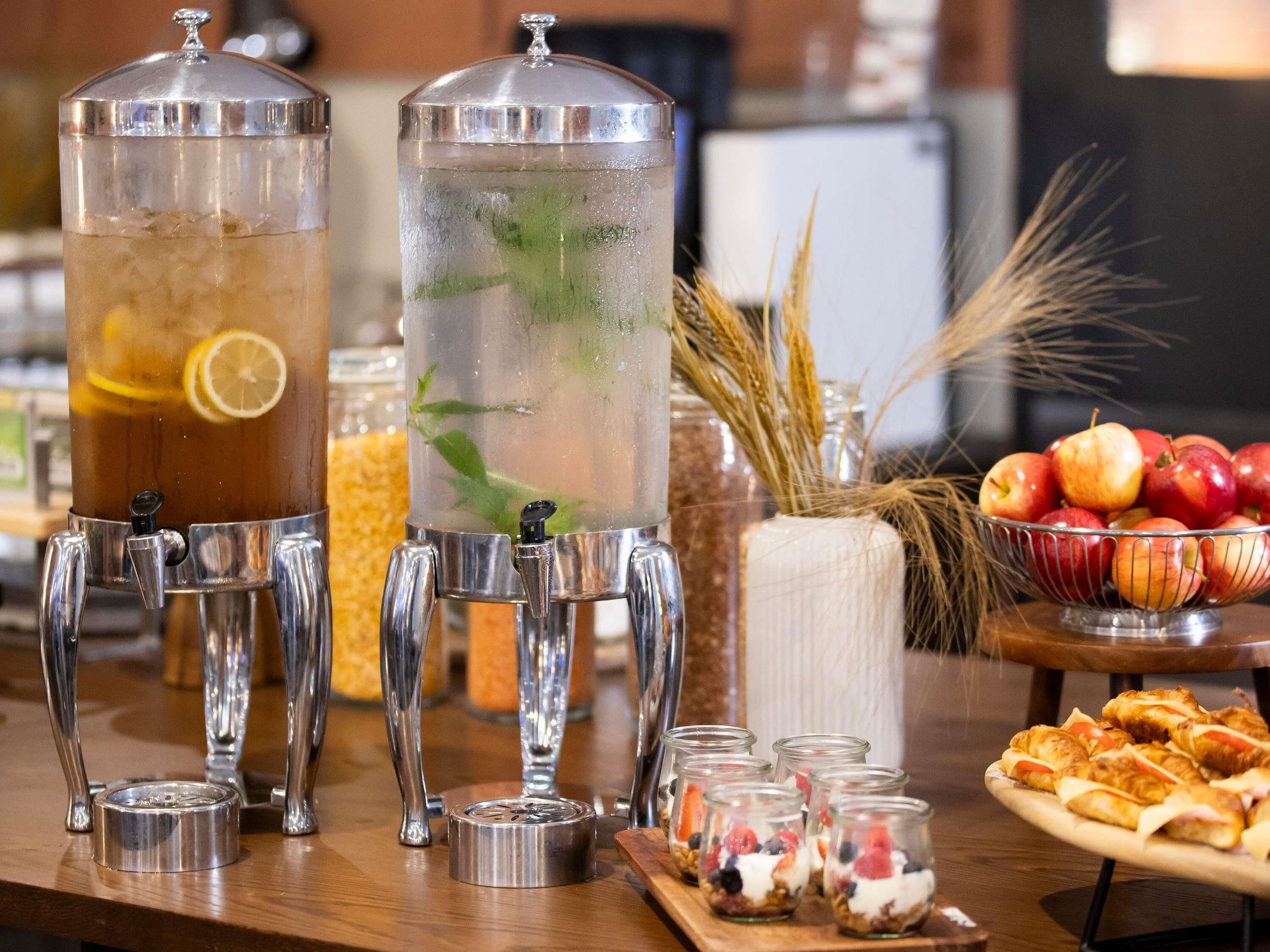 Two beverage dispensers, a bowl of apples, and a tray of pastries.