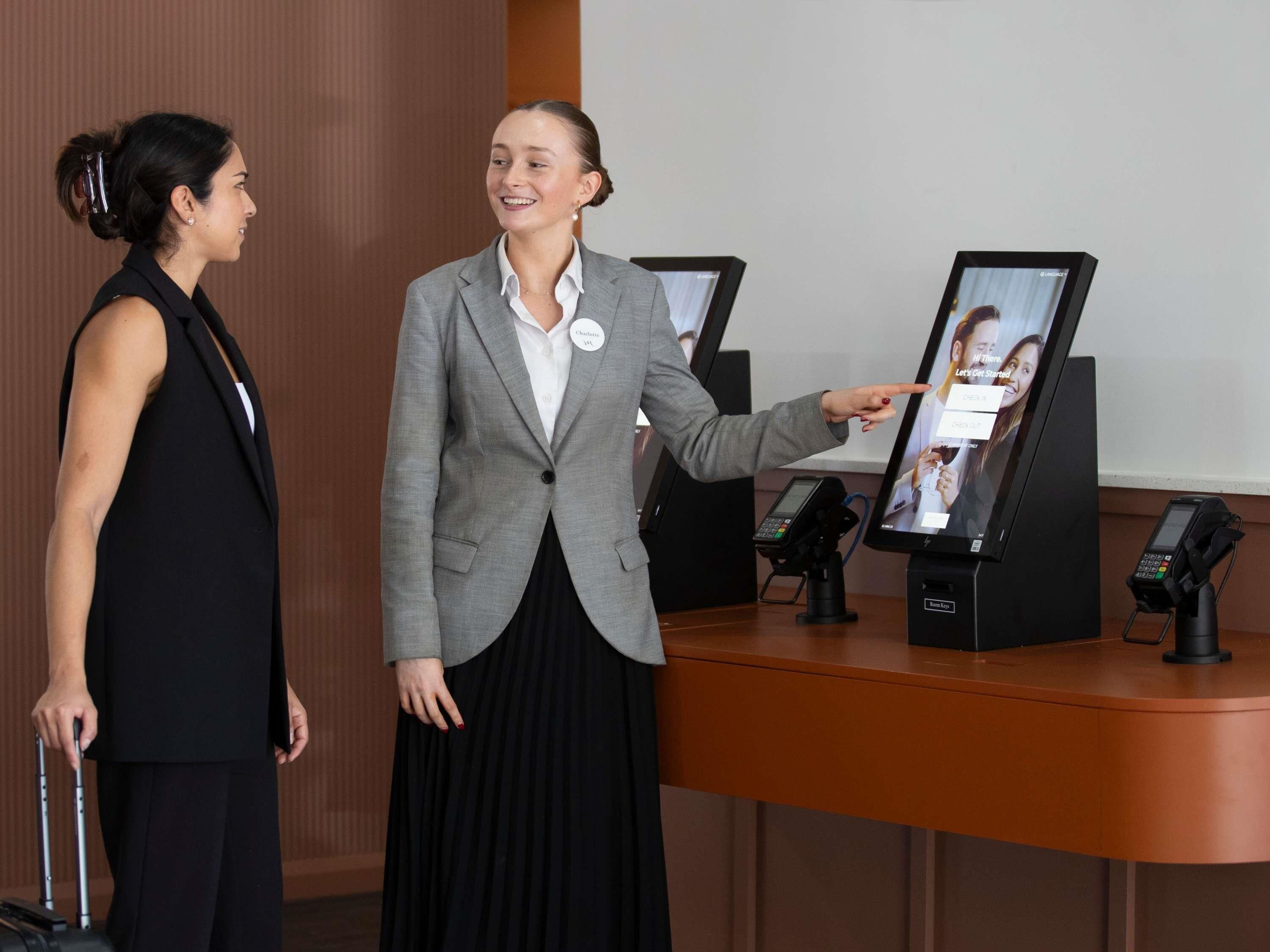 Uma área de recepção de hotel com balcão, um quiosque digital e um carrinho de bagagem.