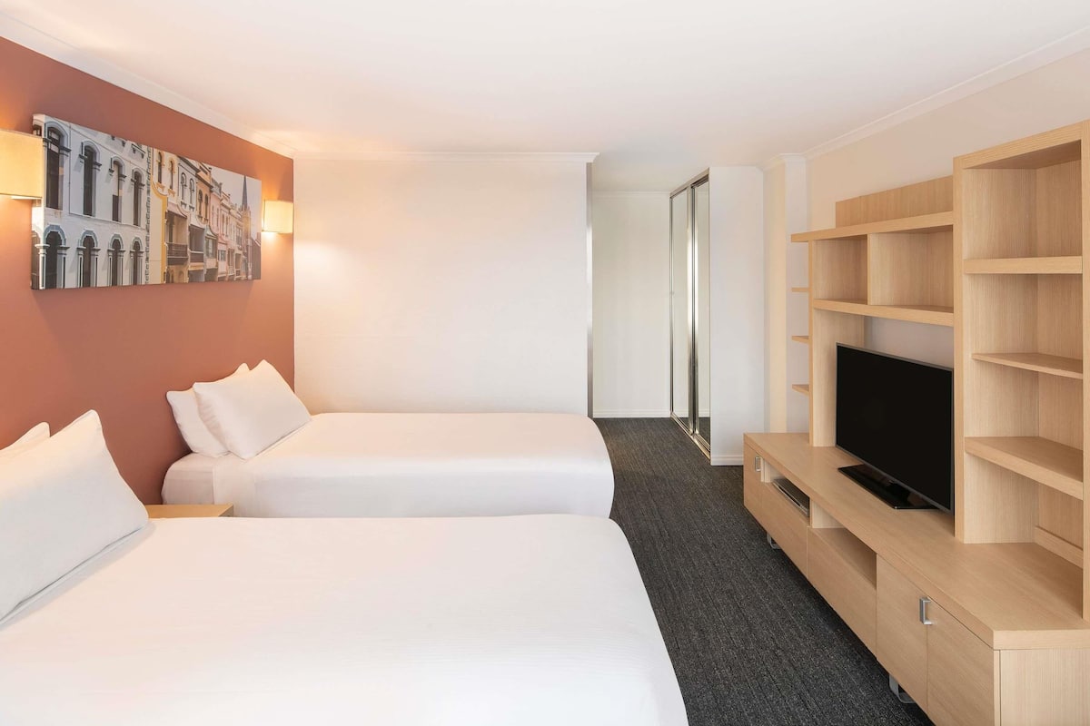 A hotel room with two beds, a TV, a wooden TV stand, a wardrobe, and a wall with a cityscape painting.
