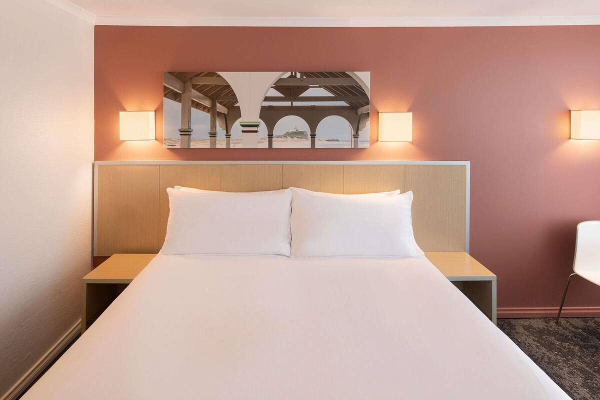 A hotel room with a bed, two bedside tables, a wall-mounted artwork, and two wall-mounted lamps.