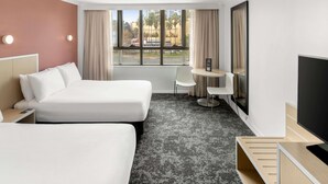 A hotel room with two beds, a desk, a chair, a TV, and a window with a view of buildings and palm trees. - Mercure Newcastle (Newcastle West)