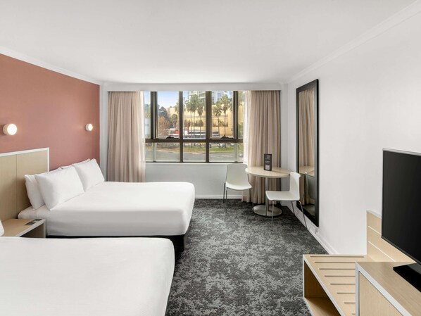 A hotel room with two beds, a desk, a chair, a TV, and a window with a view of buildings and palm trees. - Mercure Newcastle (Newcastle West)