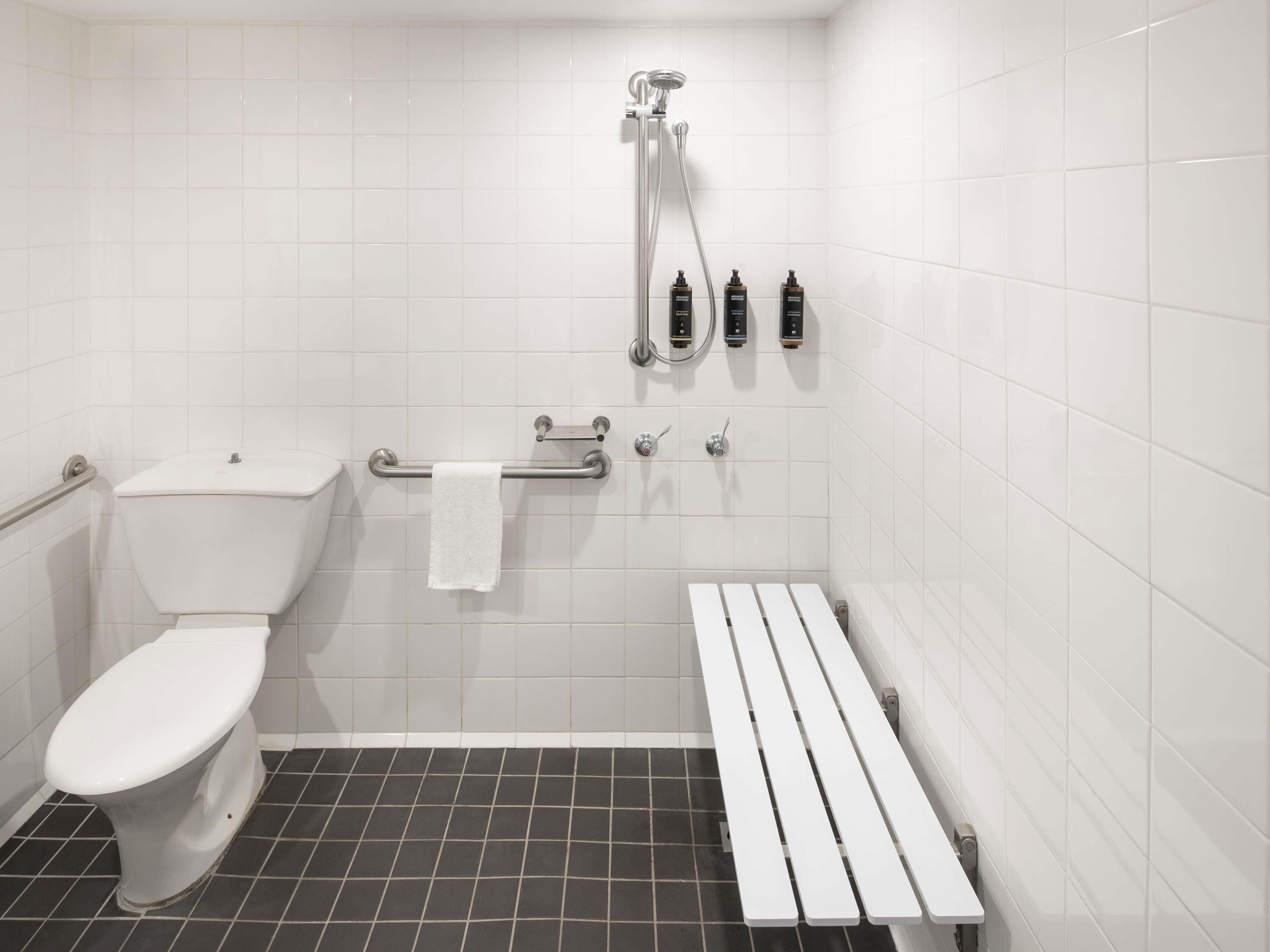 A bathroom with a toilet, a shower with a handheld showerhead, and a bench.