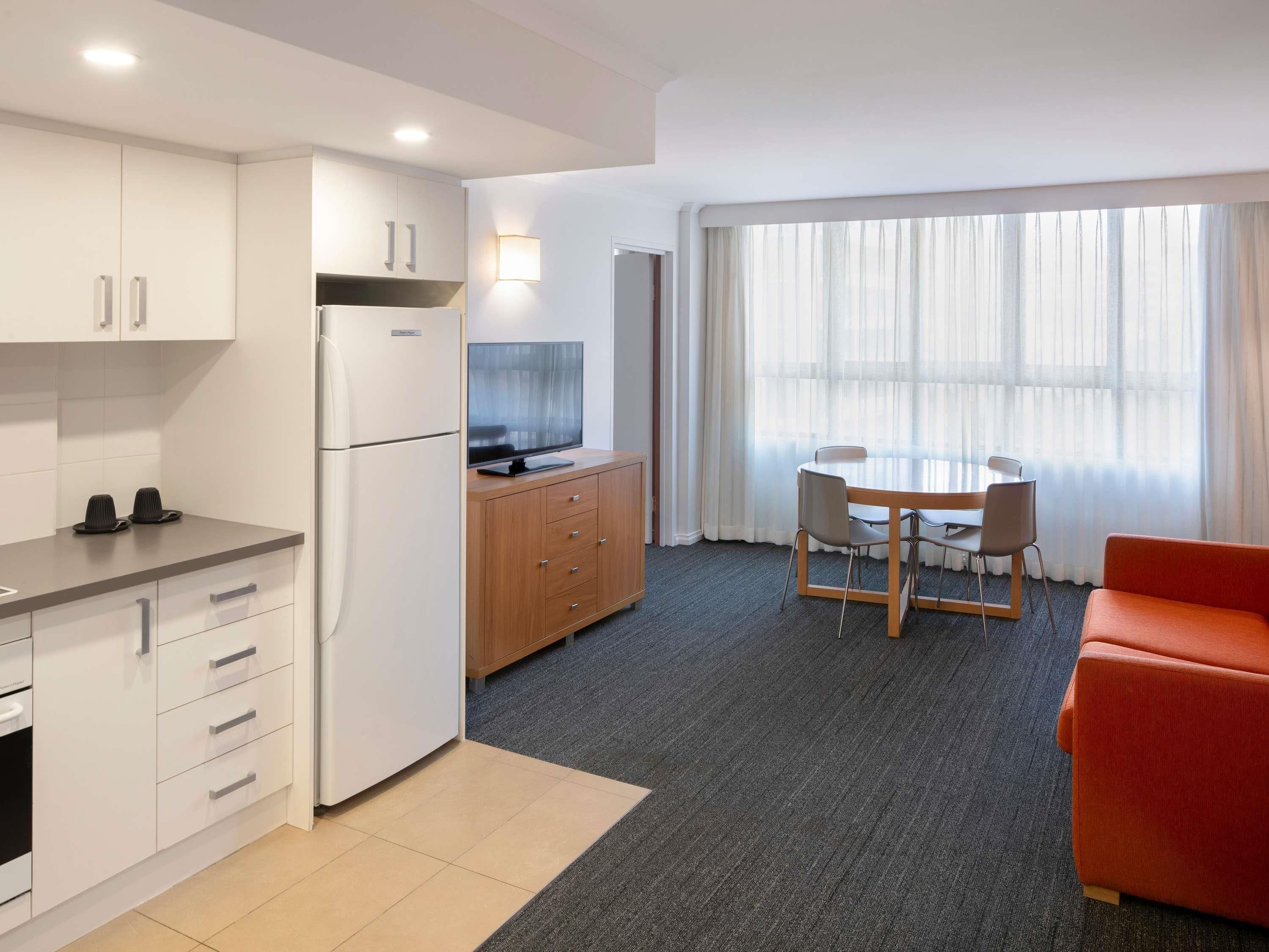 Ein modernes Hotelzimmer mit einer Küchenzeile, einem Essbereich mit Tisch und Stühlen, einem Flachbildfernseher und einem großen Fenster mit durchsichtigen Vorhängen.