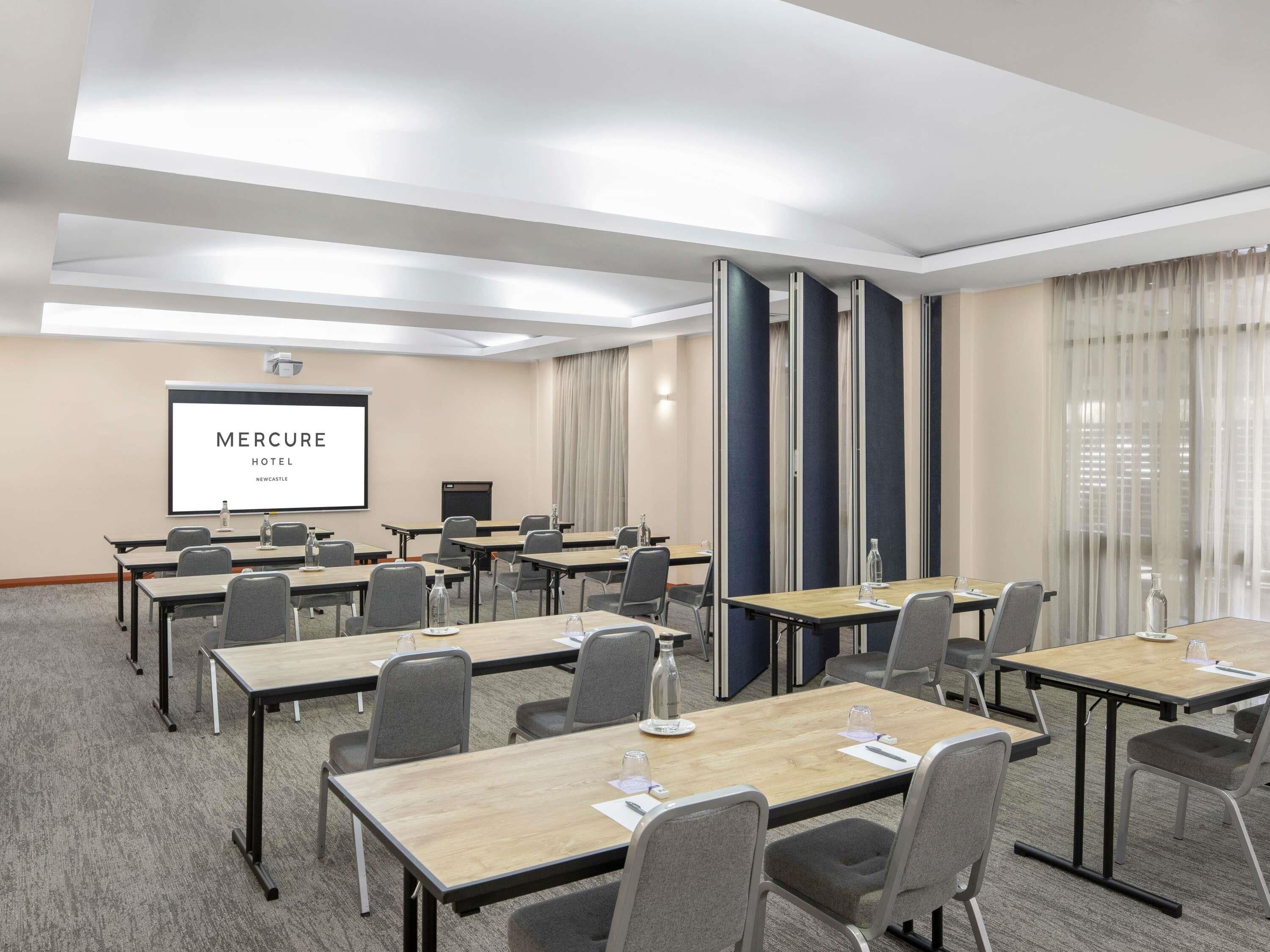 Ett konferensrum med flera bord, stolar, en projektorduk som visar texten ”MERCURE HOTEL NEWCASTLE” och ett stort fönster med gardiner.