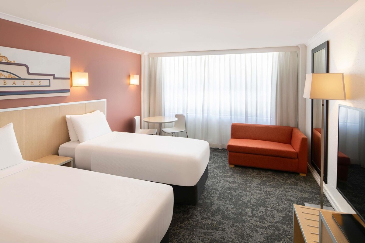 A hotel room with two beds, a small table, a sofa, a TV, and a window with curtains.
