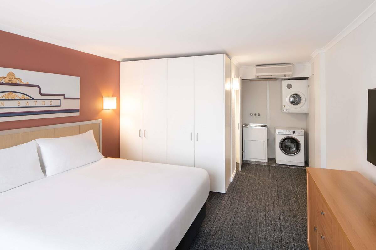 A hotel room with a bed, a wardrobe, a washing machine, and a wall-mounted TV.