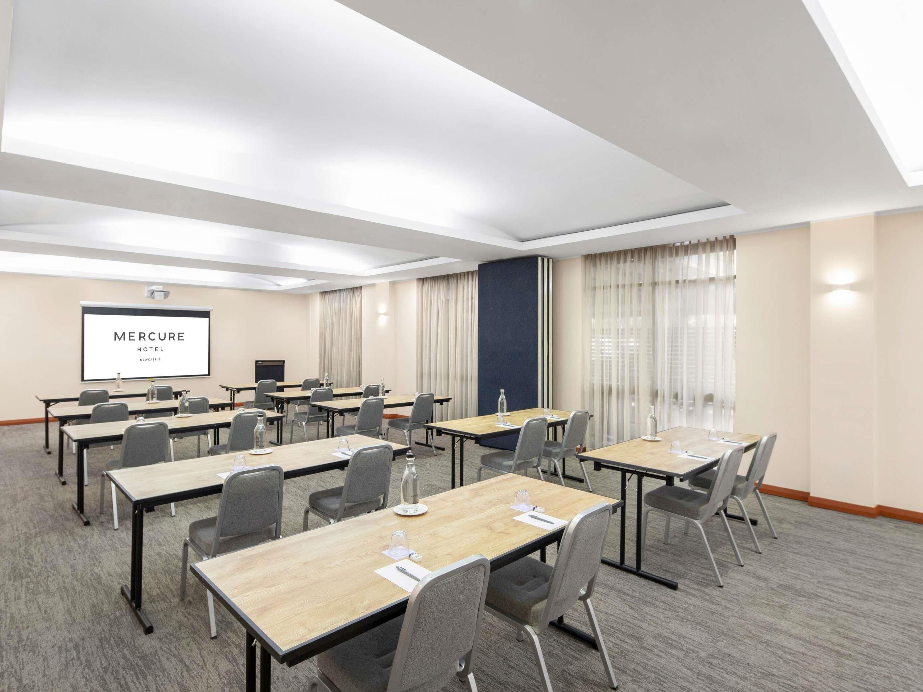 A conference room with multiple tables and chairs, a projector screen displaying 'Mercure Hotel Newcastle,' and a large window with curtains.