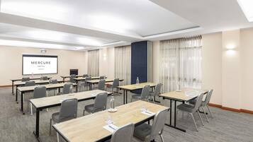 A conference room with multiple tables and chairs, a projector screen displaying 'Mercure Hotel Newcastle,' and a large window with curtains.