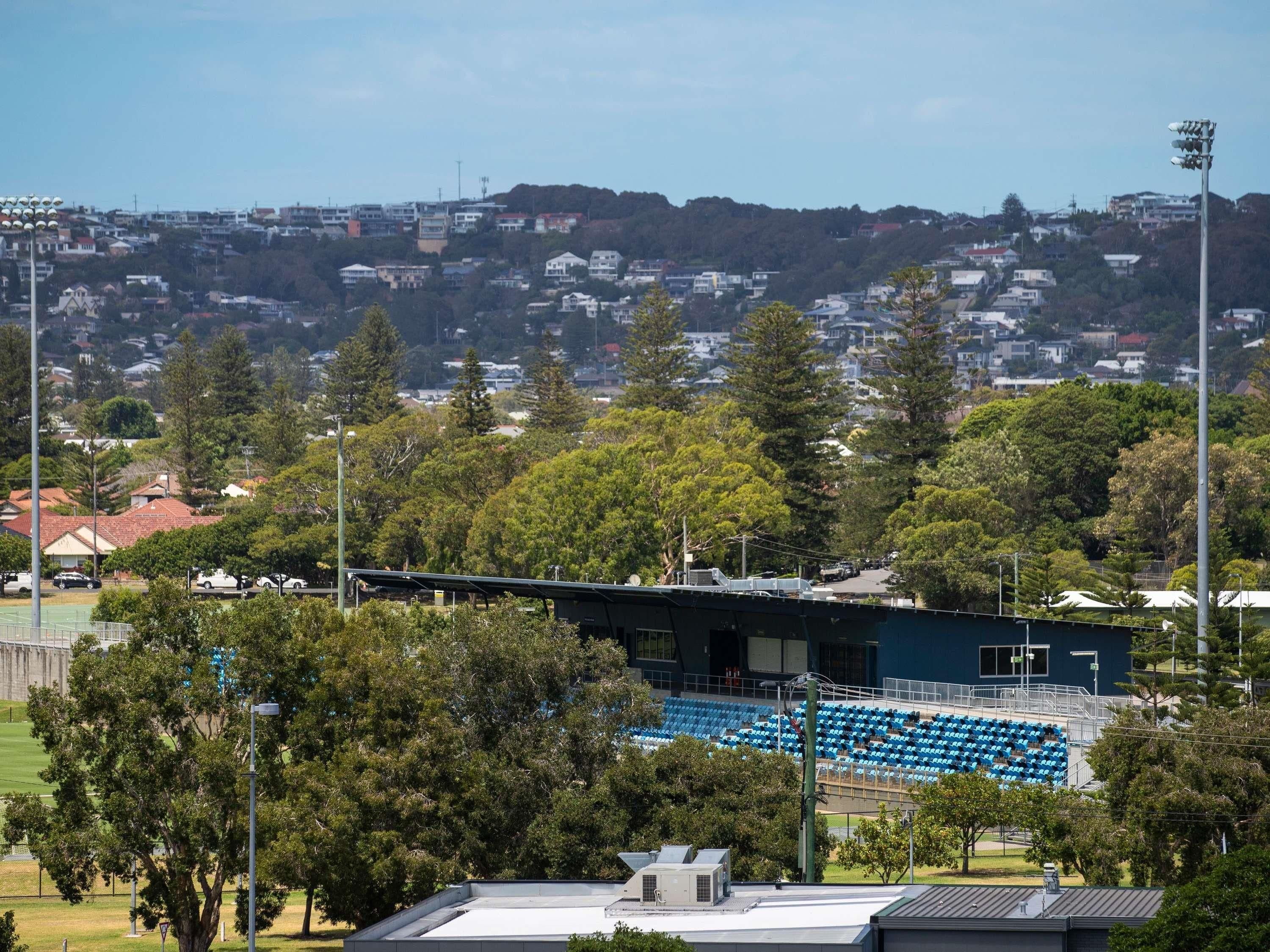 A stadium with blue seating, surrounded by trees and residential buildings in the background.