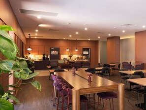 A modern dining area with wooden tables and purple bar stools, a kitchen in the background, and ceiling lights. - Mercure Newcastle (Newcastle West)