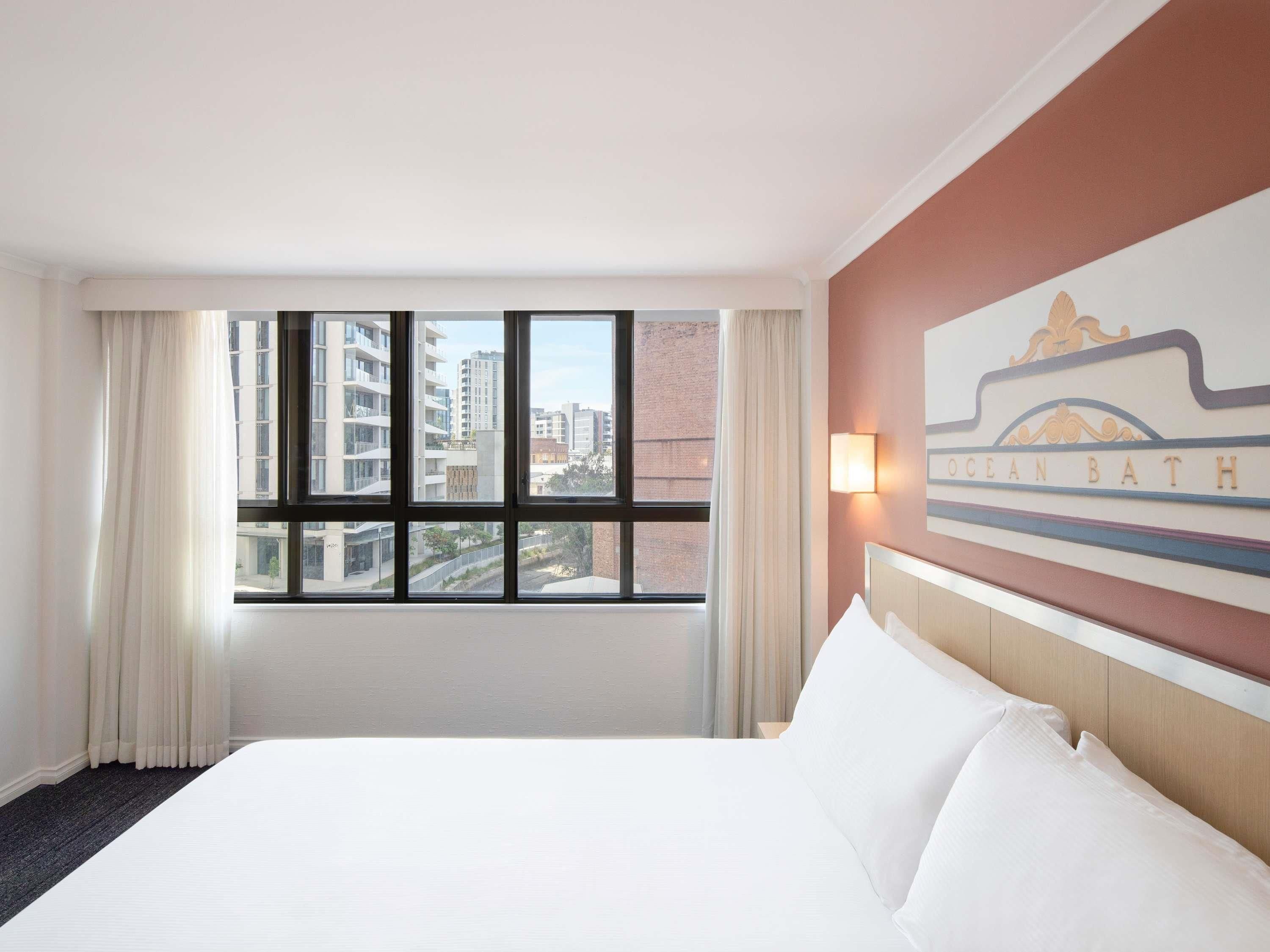A hotel room with a large bed, a window with curtains, and a wall-mounted light.