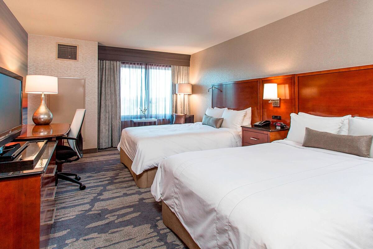 A hotel room with two beds, a desk with a computer, a chair, a lamp, and a window with curtains.