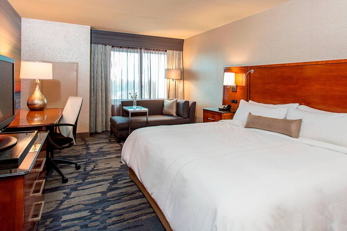A hotel room with a large bed, a desk with a lamp, a chair, a small table with a vase, and a nightstand with a phone.