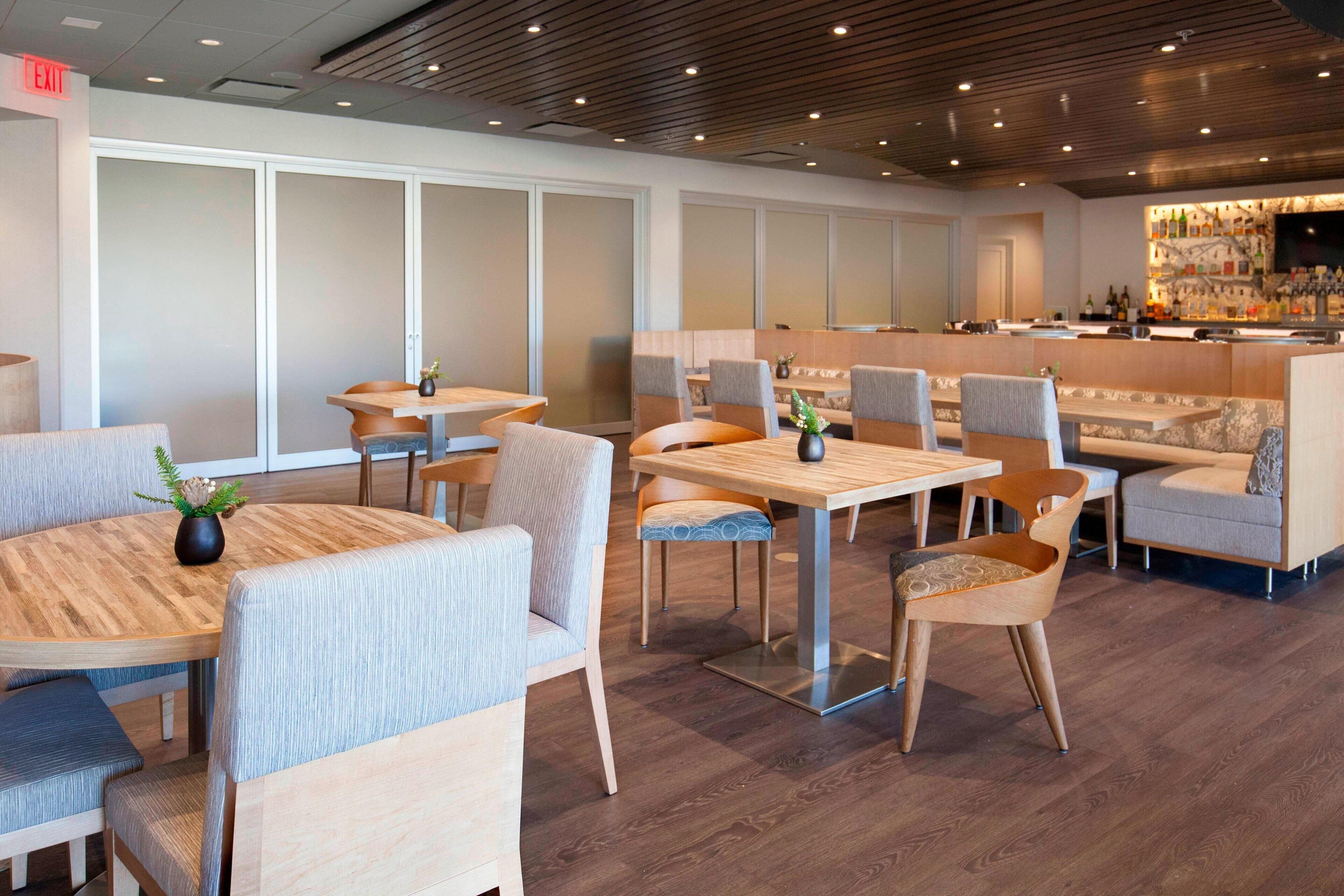 A modern restaurant with wooden tables and chairs, a bar area, and a large window with frosted glass panels.