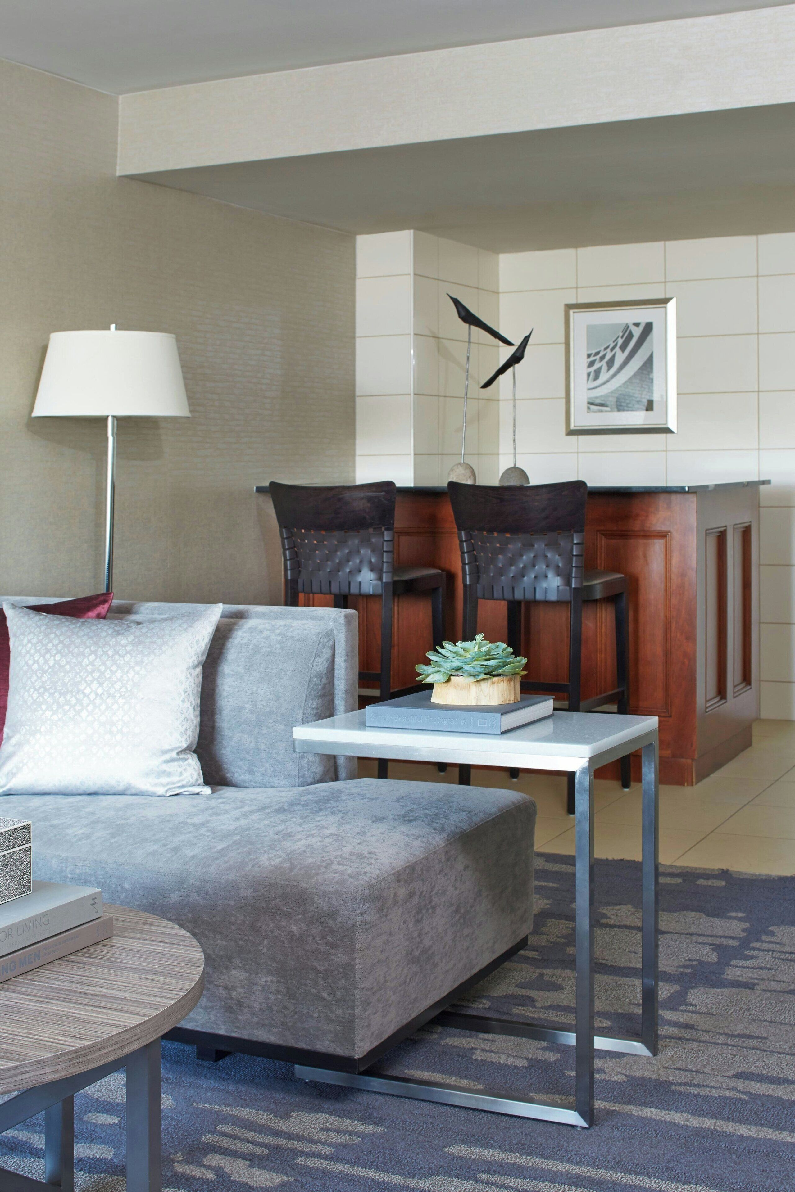 Un salon moderne avec un canapé gris, une table basse ronde et une table à manger avec des chaises. On y trouve une lampe, une plante et un tableau encadré accroché au mur.