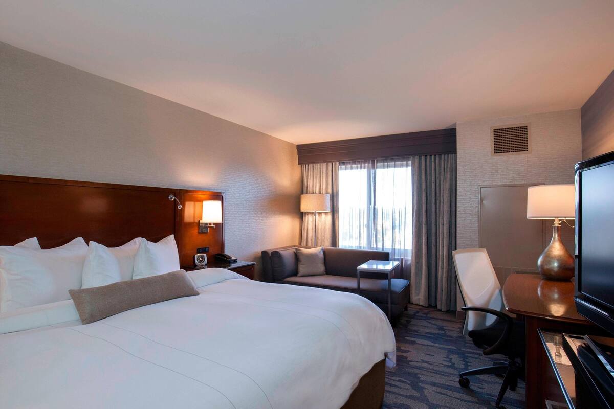 A hotel room with a large bed, a sofa, a desk with a lamp, a TV, and a window with curtains.