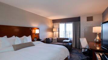 A hotel room with a large bed, a sofa, a desk with a lamp, a TV, and a window with curtains.