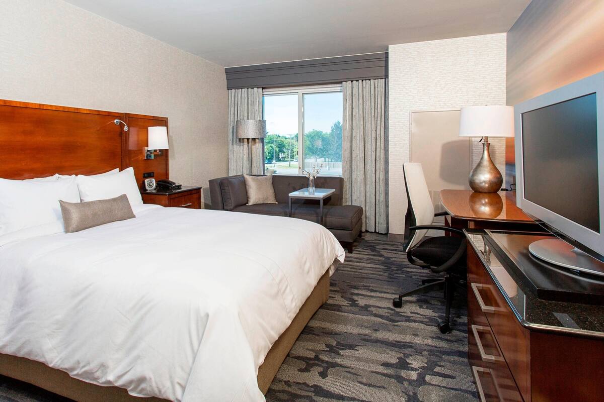A hotel room with a large bed, a desk with a TV, a chair, a small table with a lamp, and a window with curtains.