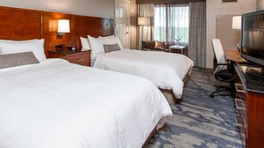 A hotel room with two beds, a desk with a television, a chair, and a small table with a lamp.