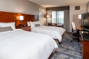 A hotel room with two beds, a desk with a television, a chair, and a small table with a lamp.