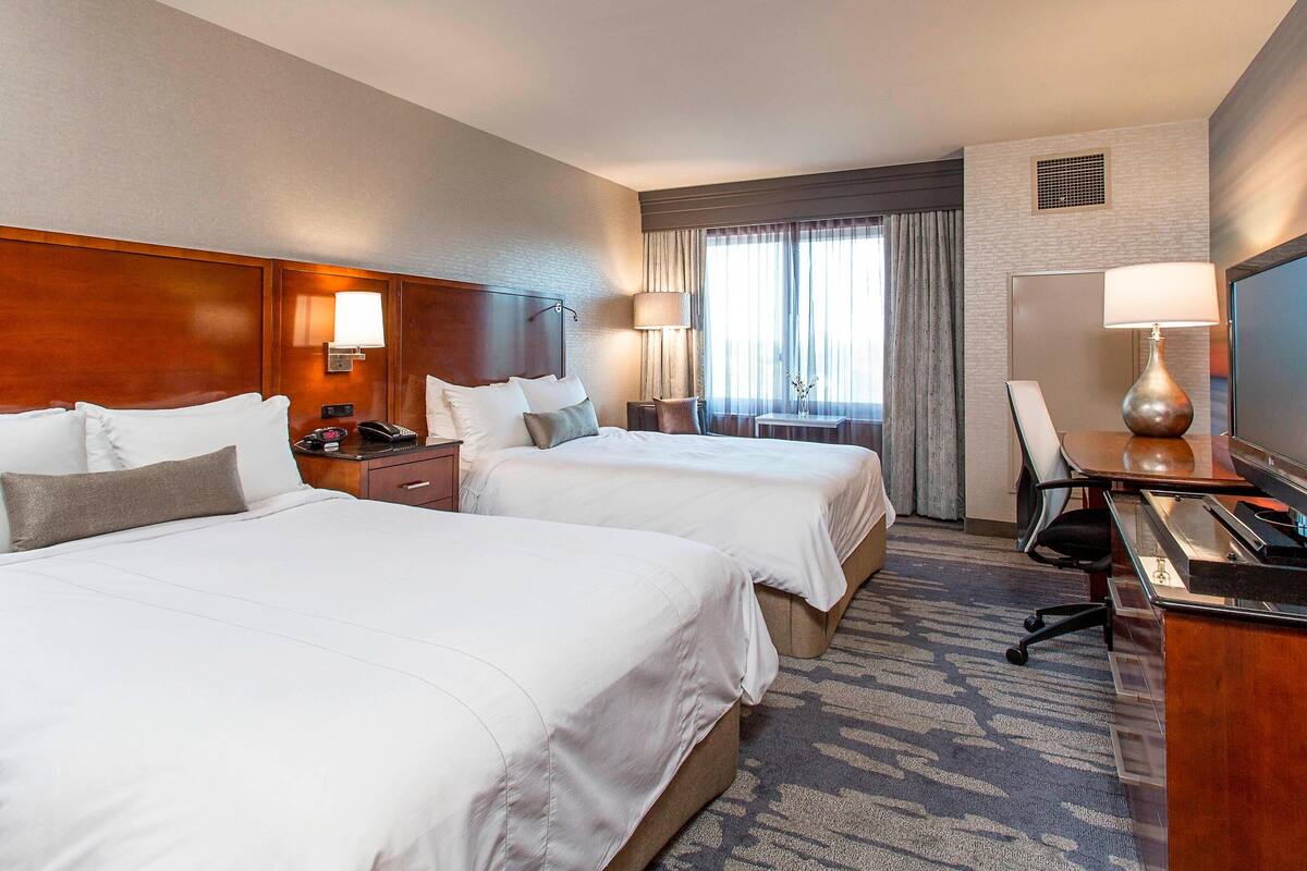 A hotel room with two beds, a desk, a chair, a TV, and a window with curtains.