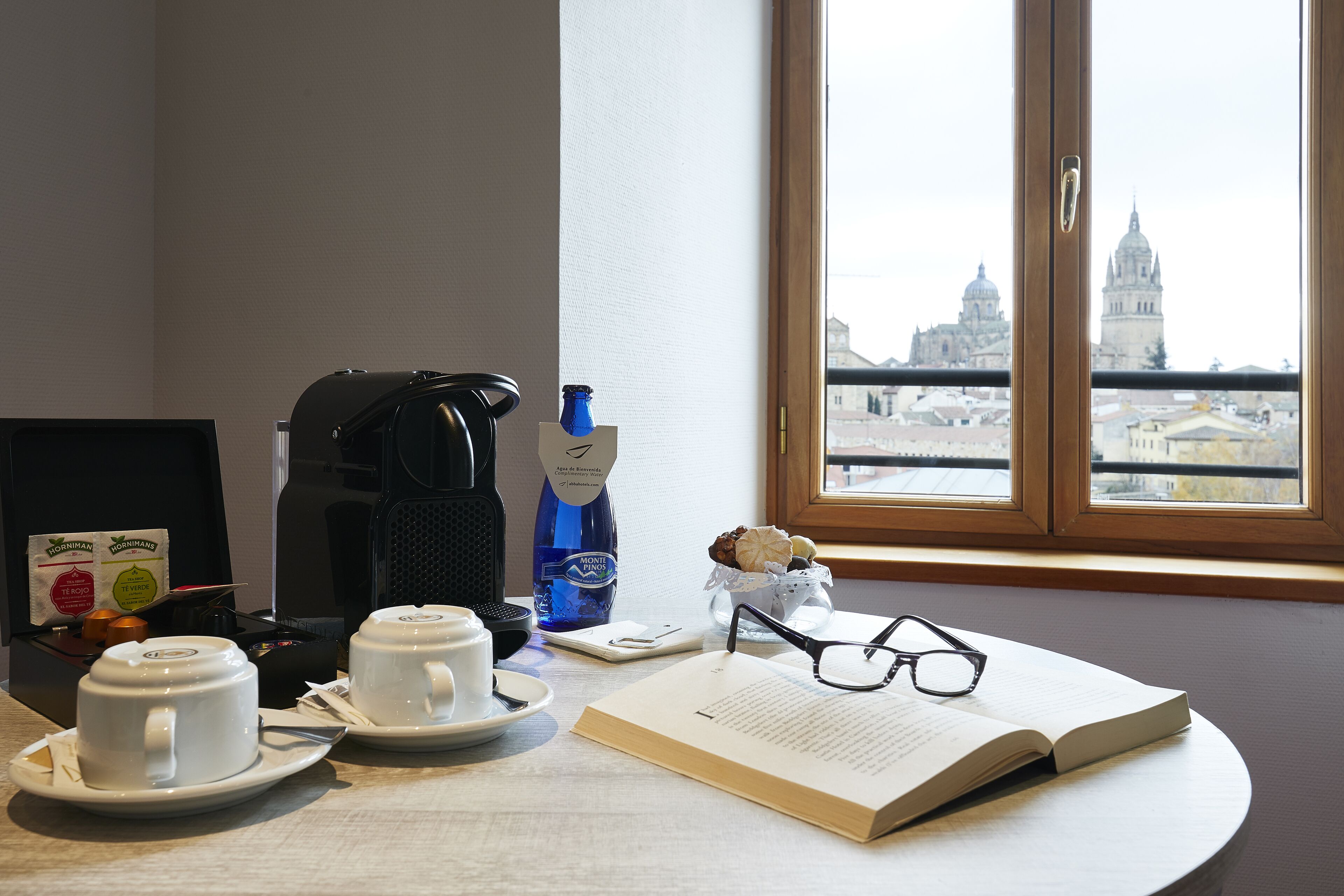 A table with two white cups, a coffee maker, a bottle of water, a small vase with nuts, a pair of glasses, and an open book.