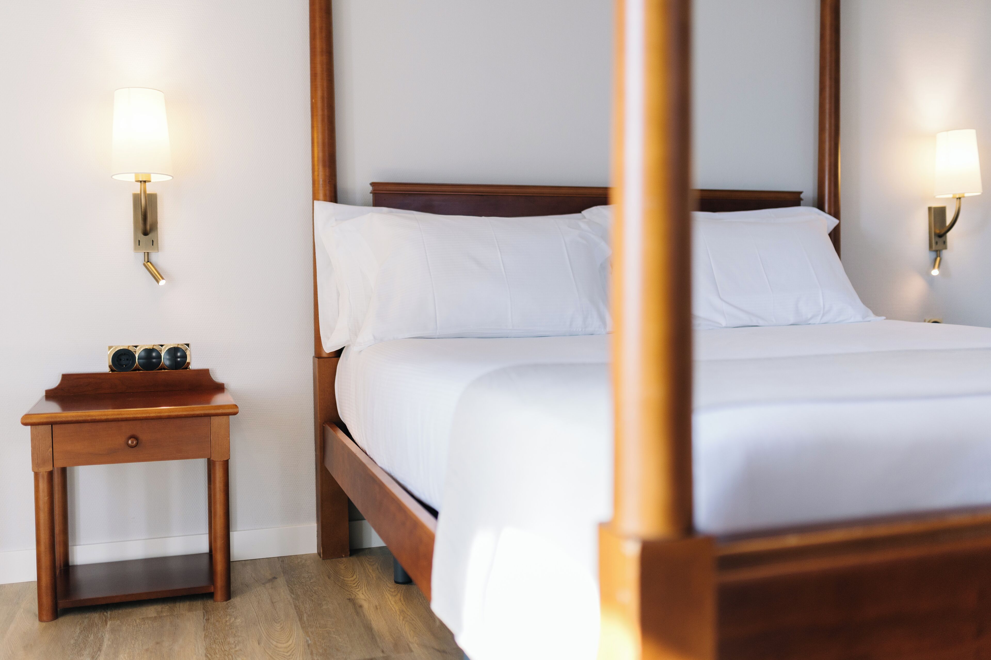 Een net opgemaakt bed met witte lakens, een houten nachtkastje met lade, en twee wandlampen aan weerszijden van het bed.