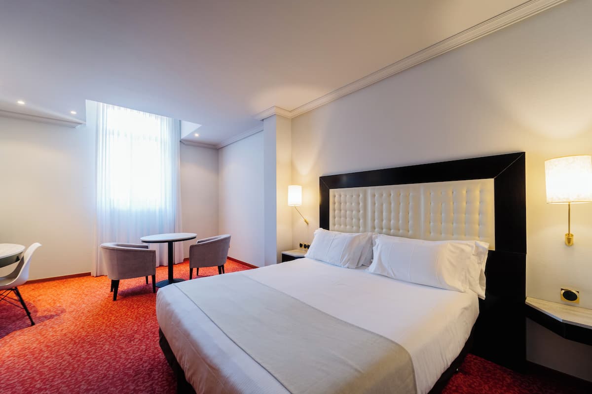 Une chambre d’hôtel avec un grand lit, une table de chevet, une chaise, une petite table et une fenêtre avec des rideaux.
