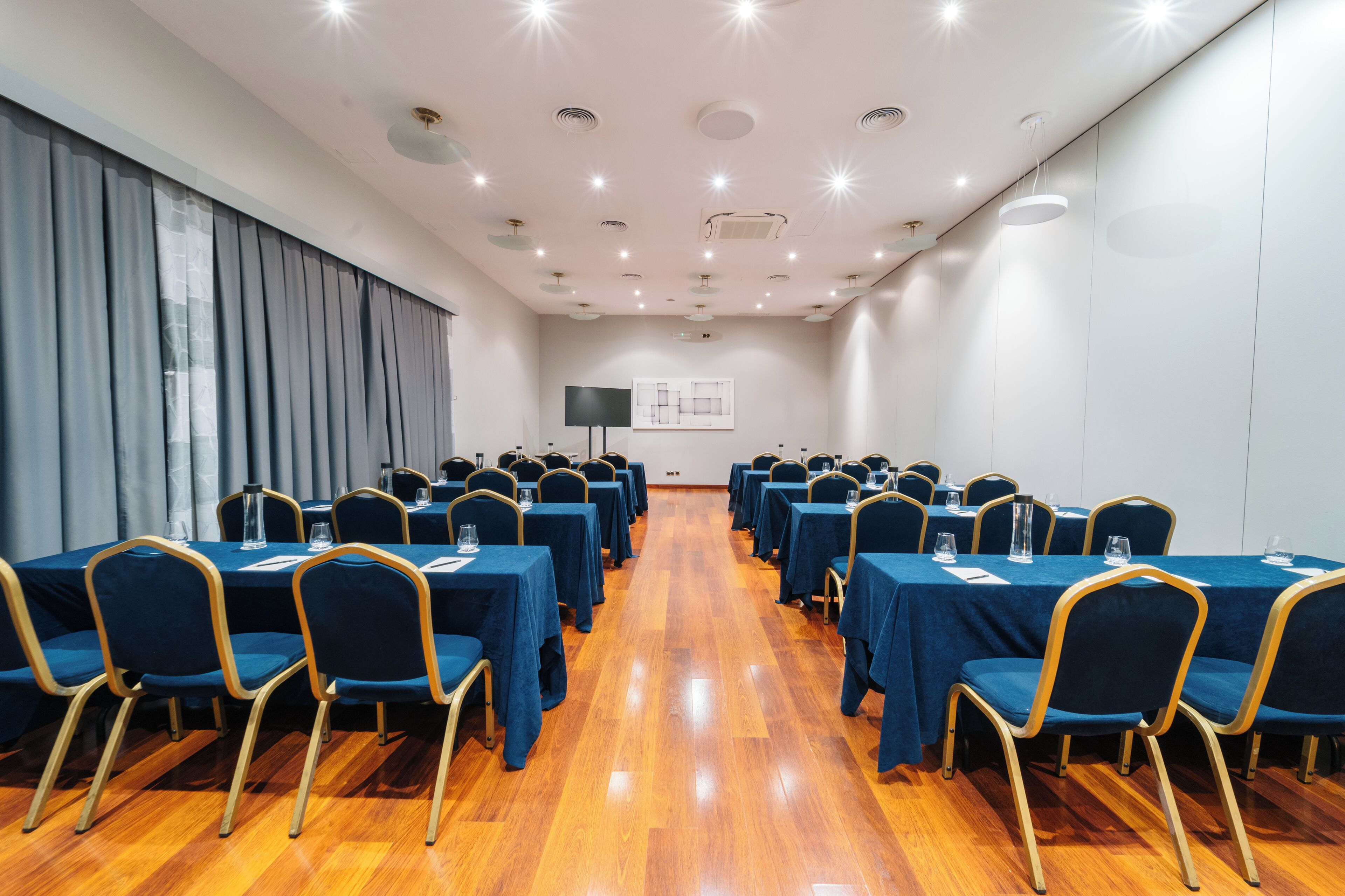 Un salón de conferencias con filas de mesas y sillas, una pantalla de proyector y un ventanal con cortinas.