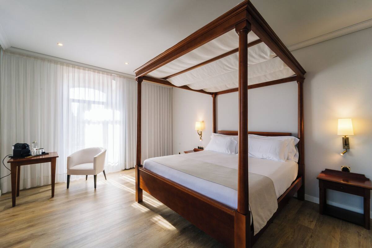 Un lit à baldaquin avec une structure en bois et une literie blanche, une table de chevet avec une lampe, une chaise et un bureau avec une lampe.