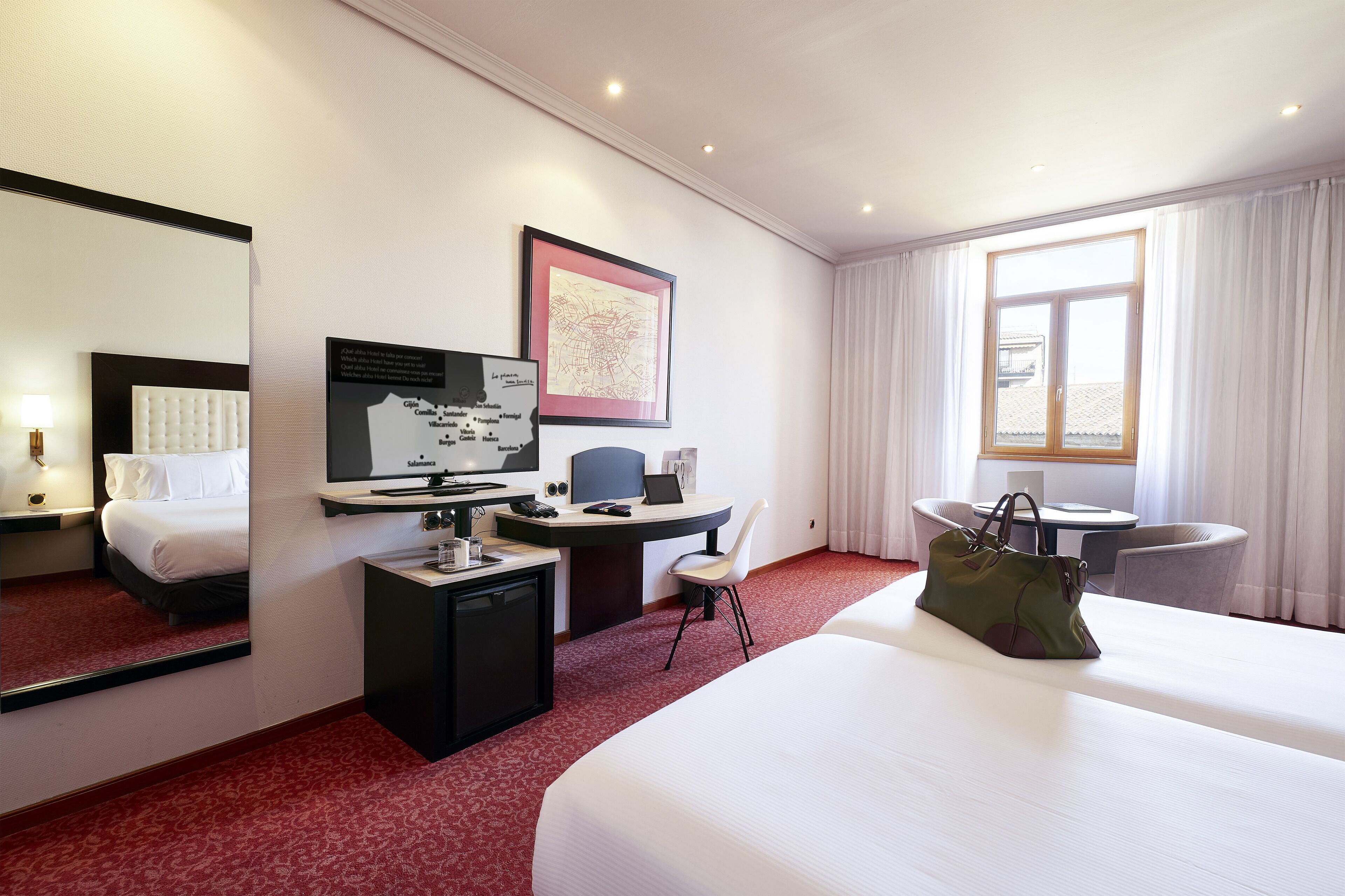 A hotel room with a large bed, a desk with a computer, a chair, a small table, a lamp, and a window with curtains.