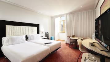 A hotel room with two beds, a desk, a TV, and a view of the city through the window.