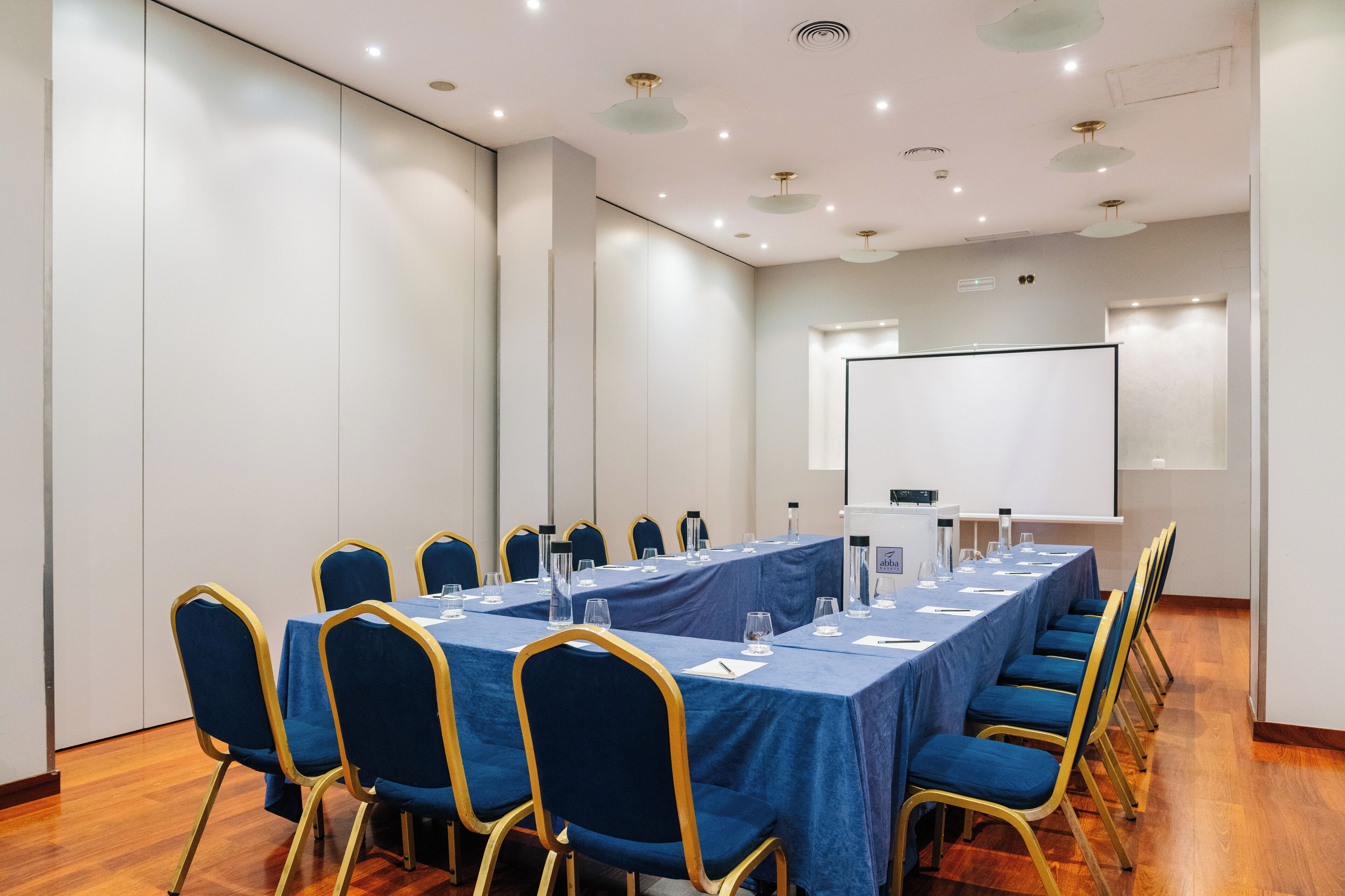 Un salón de conferencias con una larga mesa, sillas azules y una pantalla para proyector.
