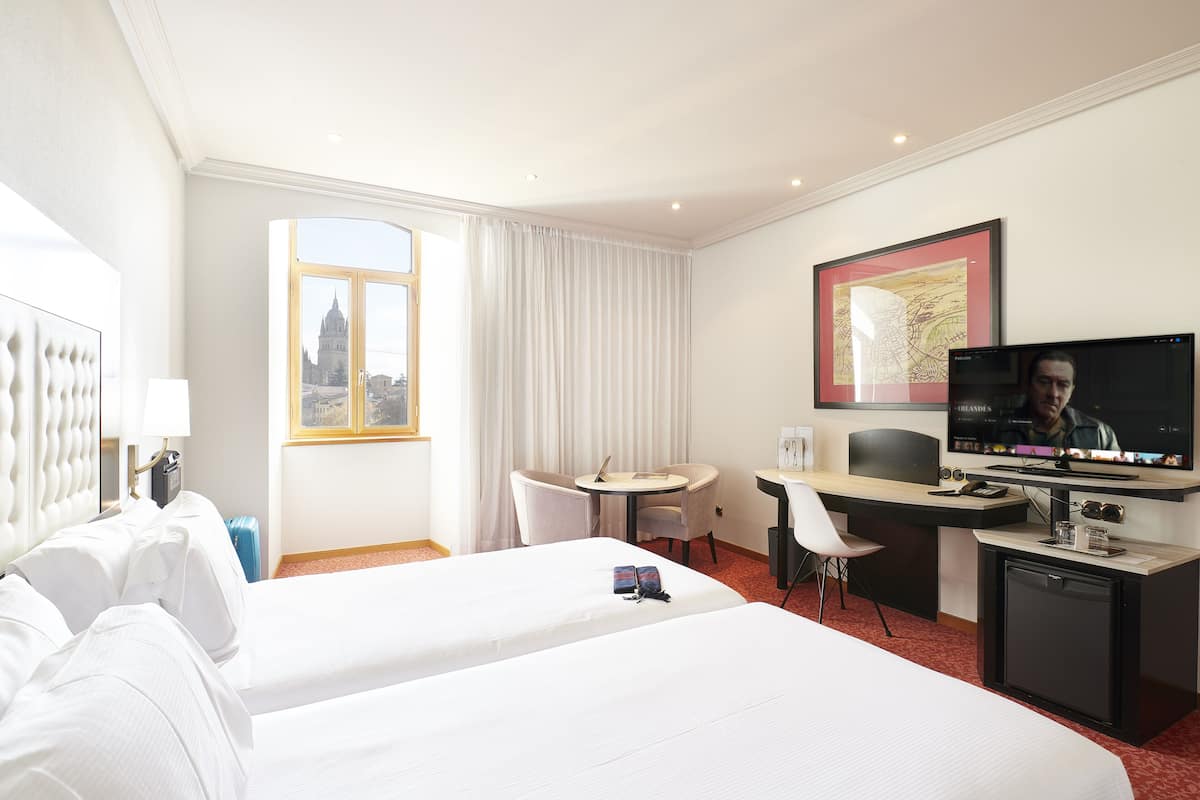 Une chambre d’hôtel avec deux lits, un bureau, une chaise, une télévision et une fenêtre donnant sur la ville.