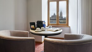 Habitación de hotel con una mesa redonda, dos sillones, una cafetera, una botella de agua y un libro. Una ventana ofrece vistas de un edificio.
