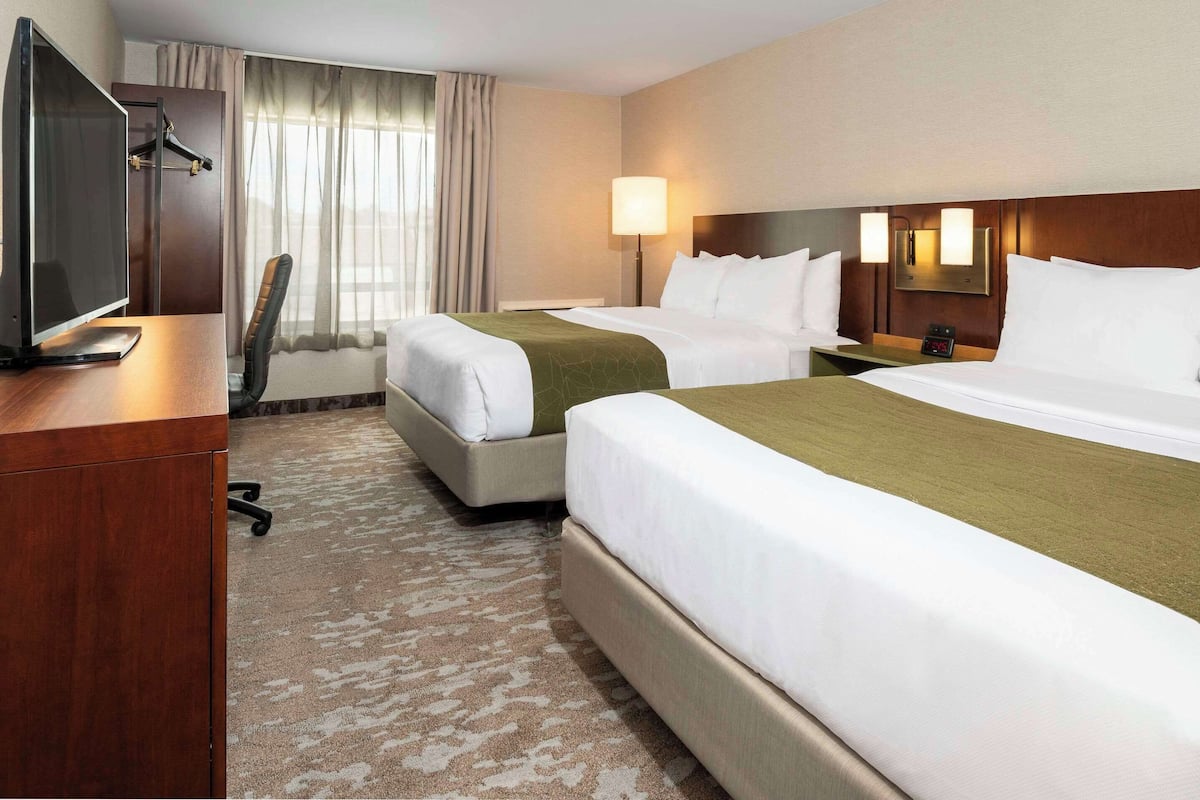 A hotel room with two beds, a desk with a computer, a chair, a lamp, and a window with curtains.