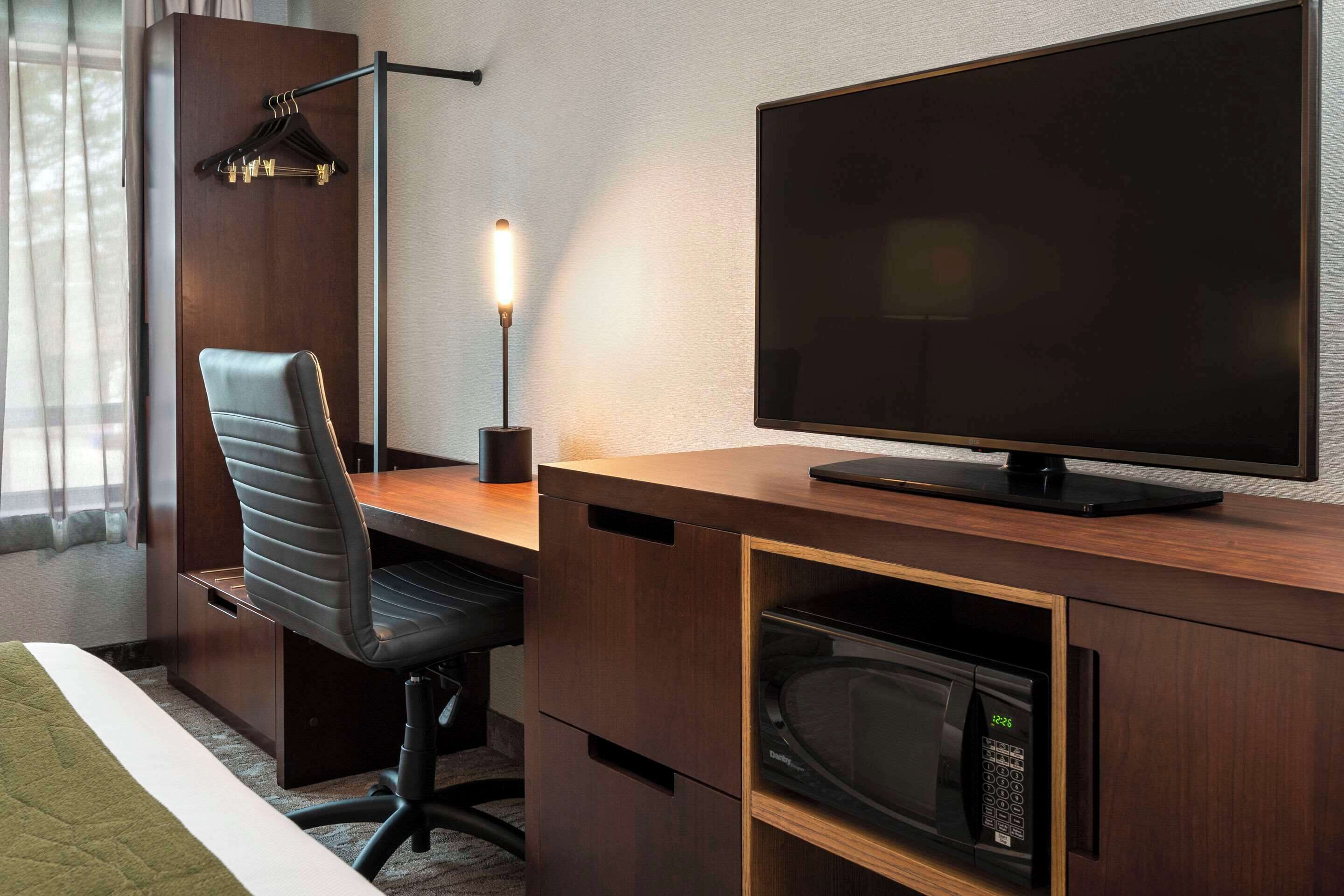 A hotel room with a wooden desk, a black leather chair, a flat-screen TV, a desk lamp, and a microwave.