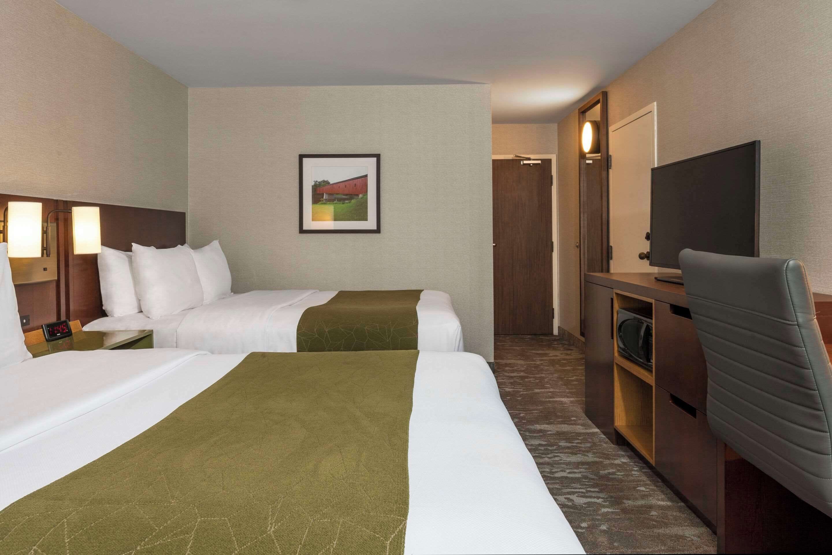 A hotel room with two beds, a TV, a desk with a lamp, and a framed picture on the wall.