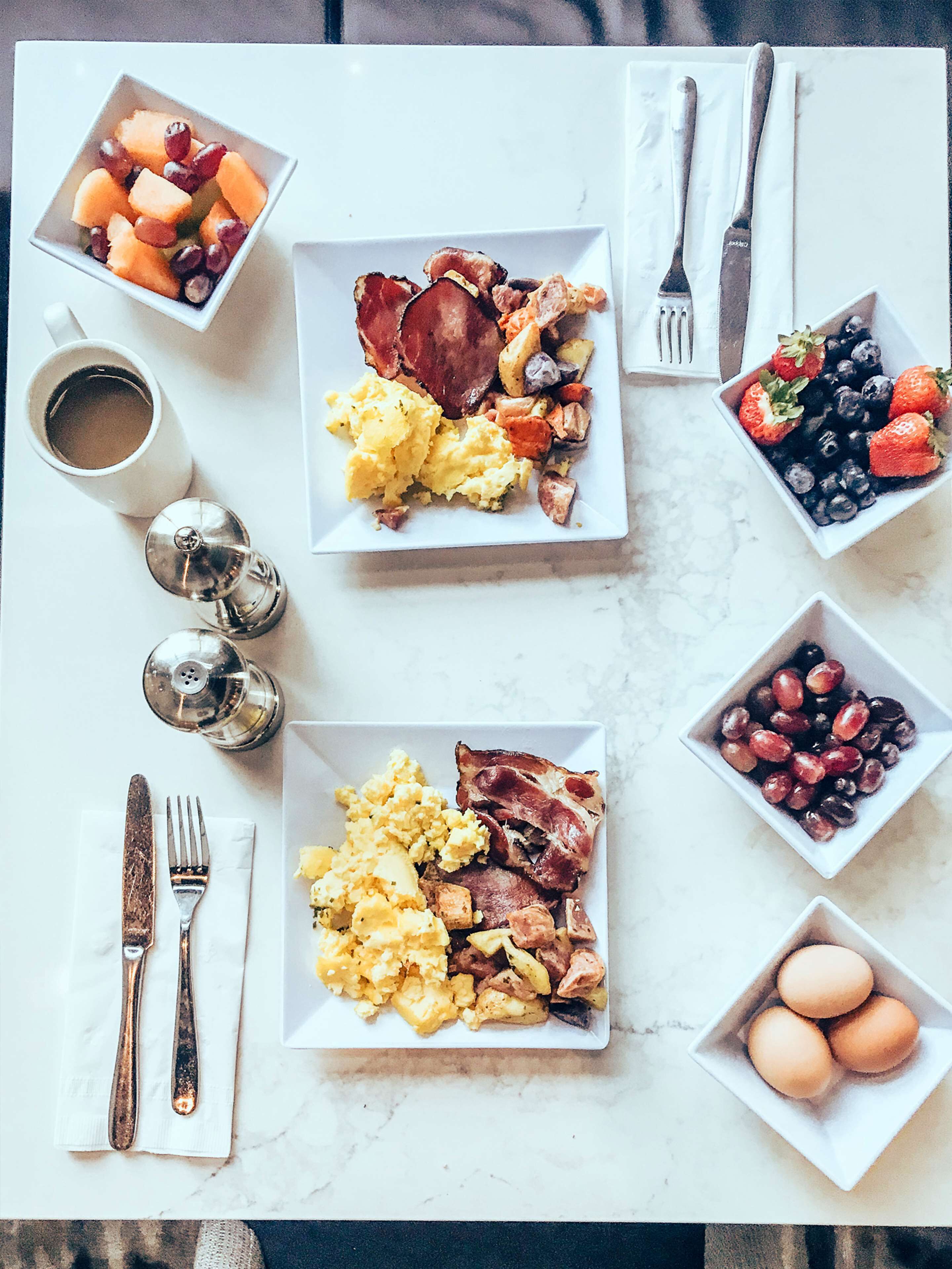 A breakfast table with plates of scrambled eggs, bacon, and toast, bowls of fruit and berries, and a cup of coffee.