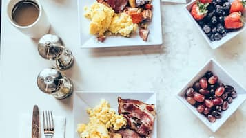 A breakfast table with plates of scrambled eggs, bacon, and toast, bowls of fruit and berries, and a cup of coffee.