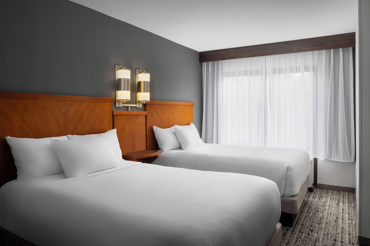 Ein Hotelzimmer mit zwei Betten, einem Holzkopfende, einem Nachttisch und einem Fenster mit durchsichtigen Vorhängen.