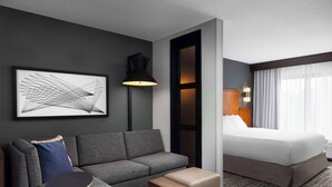 Habitación de hotel con un sofá gris, una pequeña mesa de madera, una cama con ropa blanca, una lámpara de pie negra y una obra de arte abstracto enmarcada en la pared.