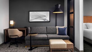Un hotel moderno con un sofá gris, una mesita redonda pequeña, un escritorio con una silla, una lámpara de pie y una gran obra de arte abstracta en la pared.