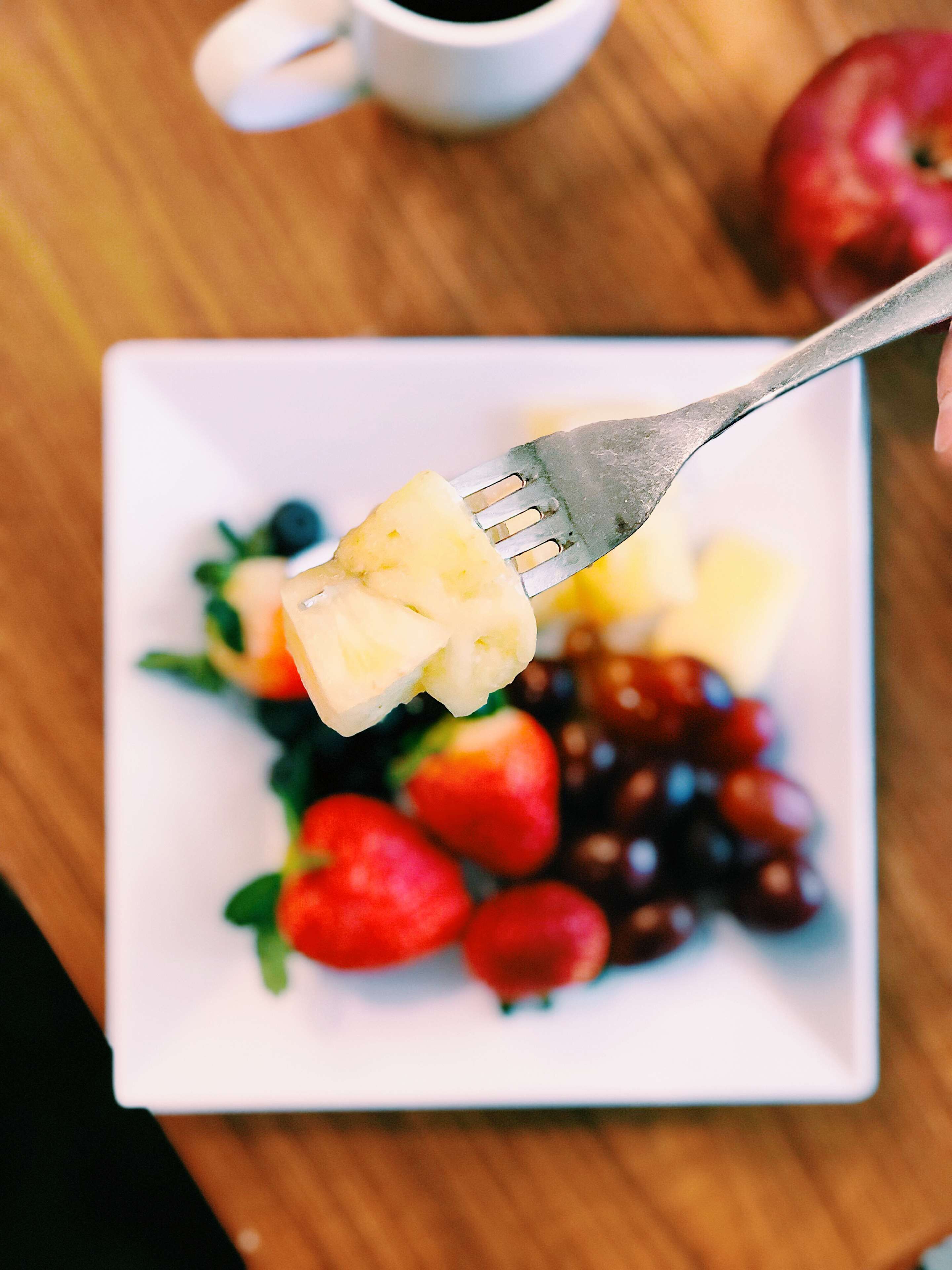 Um prato branco com um garfo segurando uma fruta, rodeado de morangos e uvas, sobre uma mesa de madeira.