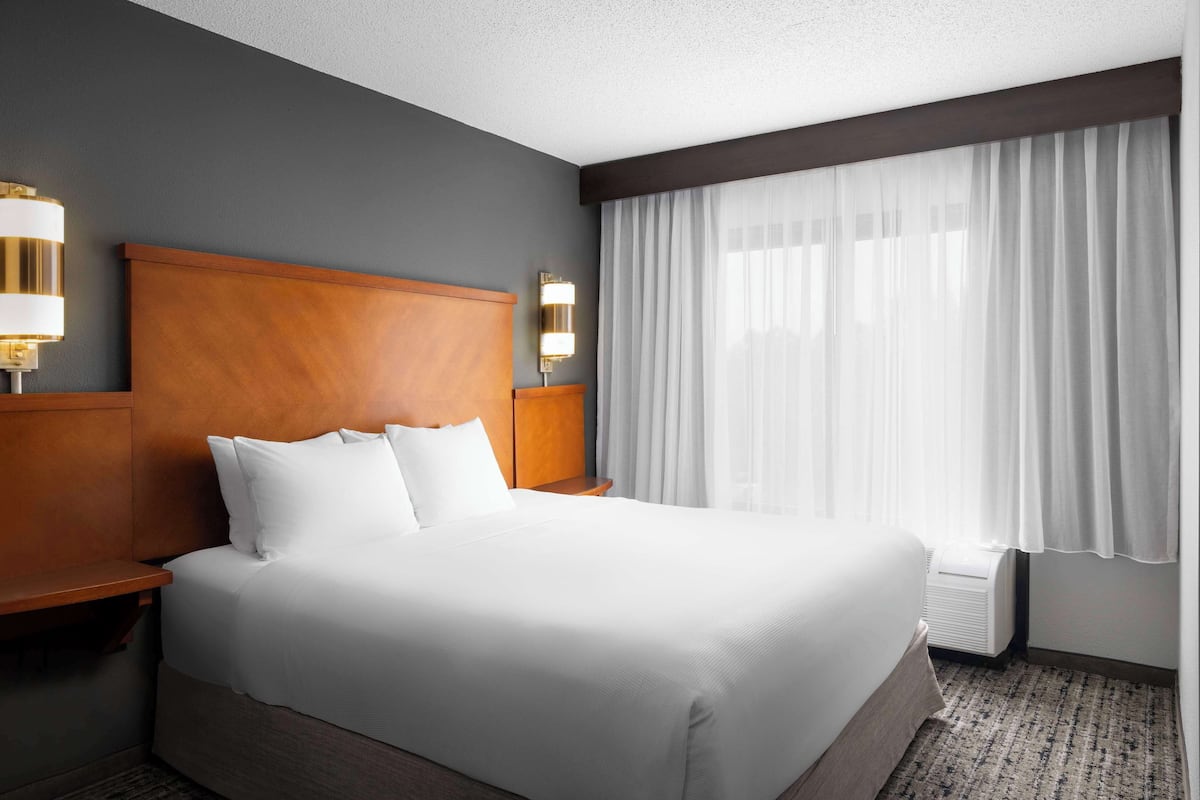 Ein Hotelzimmer mit einem großen Bett, einem Holzkopfteil, zwei Nachttischen mit Lampen, einem großen Fenster mit durchsichtigen Vorhängen und einem Teppichboden.